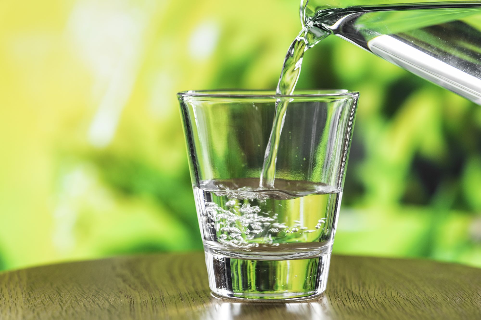 Glass with water being poured into it on a table with greenery in background